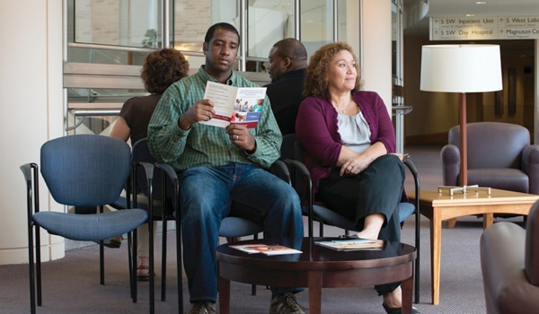 Image of a young man and a middle-aged woman sitting in a waiting room. The man is reading a pamphlet and the woman has her legs crossed and looking up.