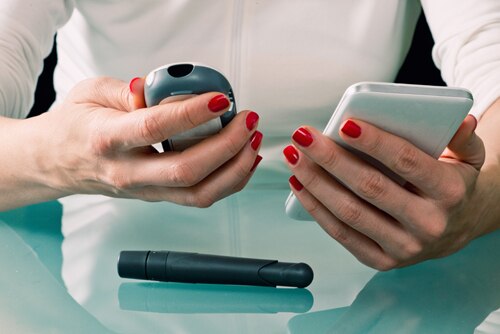 A woman holding a blood glucose test meter and a smartphone used to log results. On the table is a finger-prick tool, called a lancet, which is used to draw a drop of blood.