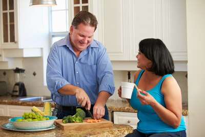 A man chopping vegetables in the kitchen and a woman sitting on a stool talking with him.