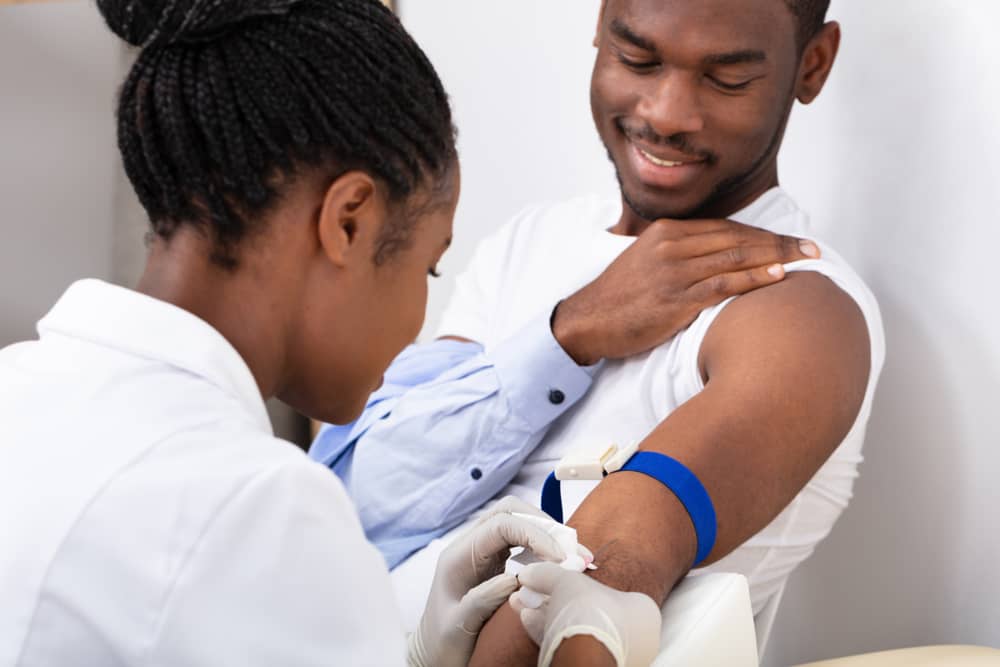 A Black health care professional taking blood from the arm of a Black male patient.