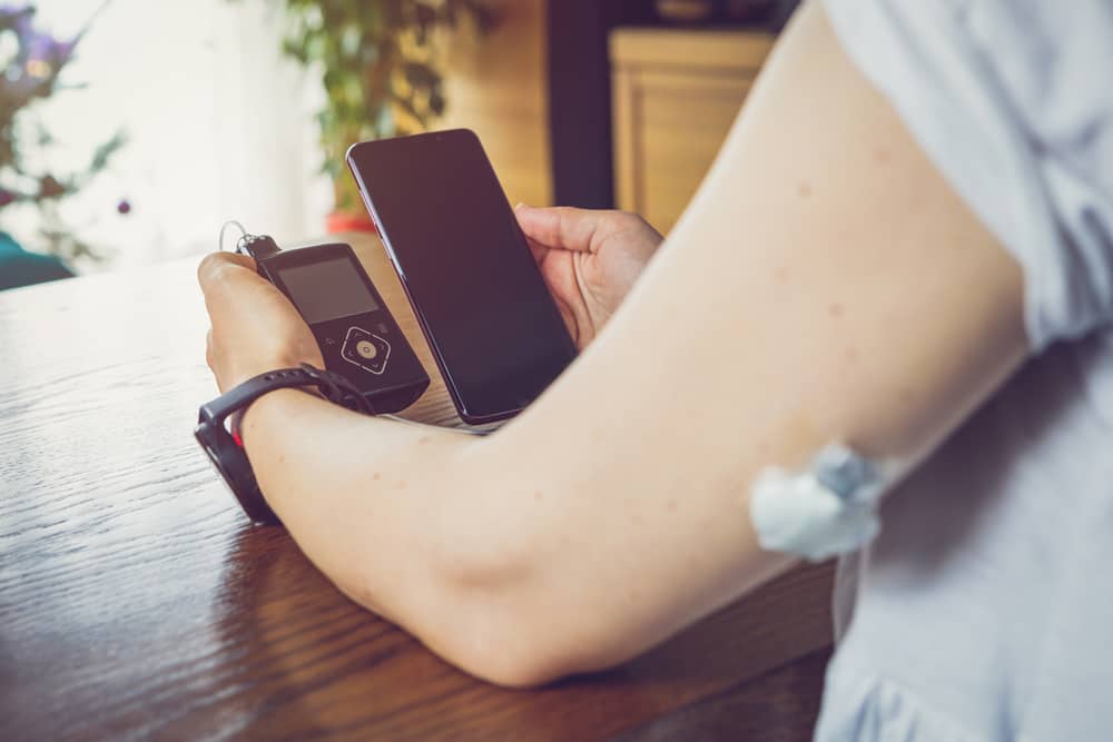 A woman is seated at a table holding a smartphone and a continuous glucose monitor, which is connected to a needle inserted at the back of her upper arm.