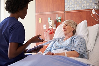 Una profesional de atención médica hablando con una paciente mayor recostada en una cama de hospital.