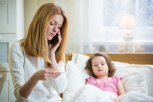 A mother holding a thermometer and sitting with a sick child while calling a doctor on the phone.