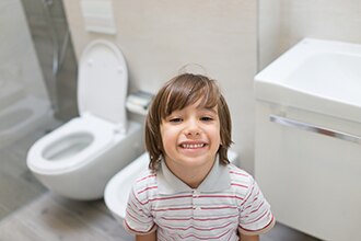 A boy smiles in a bathroom with an open toilet in the background.