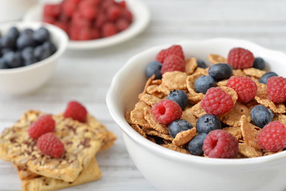 A bowl of whole-grain cereal with fresh berries next to two high-fiber crackers and raspberries.