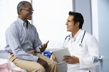 Male doctor talking to male patient sitting on an examining table.