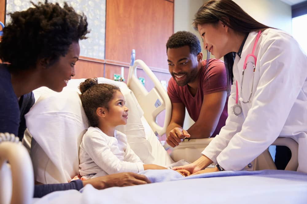 Una niña sentada en una cama de hospital, rodeada por sus padres y su doctora.
