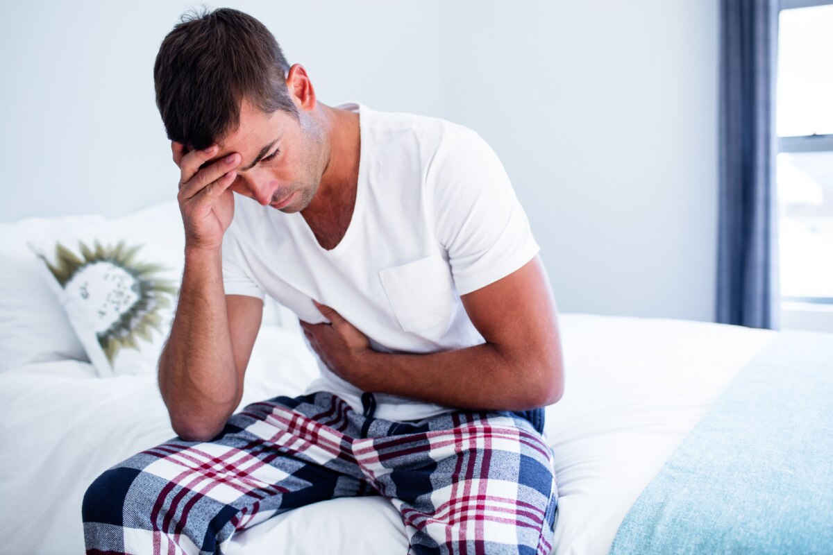 Man holding his stomach and head while sitting on a bed.