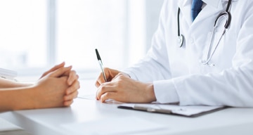 Un médico escribe una nota en un escritorio frente a un paciente, que tiene las manos cruzadas sobre el escritorio.