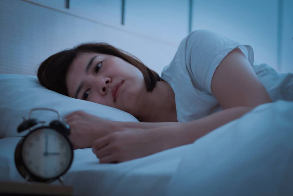 A woman lies awake in bed, facing a clock that reads 3 o’clock.