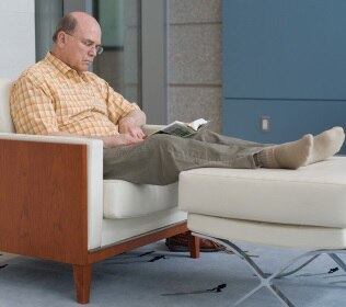 Un hombre sentado lee un libro y descansa los pies en un pequeño sillón para los pies.