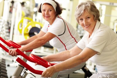Dos mujeres de mediana edad que sonríen mientras hacen ejercicio en bicicletas estacionarias.