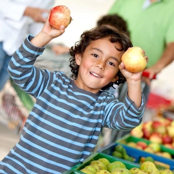 Niño parada al lado de la sección de frutas de un supermercado con una manzana en cada mano.