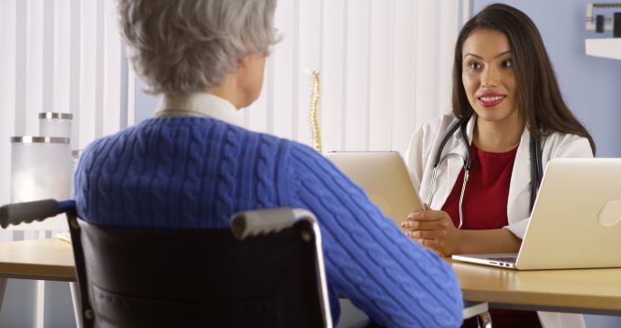 Una profesional de la salud sentada detrás de su escritorio habla con una paciente sentada frente a ella en una silla de ruedas.