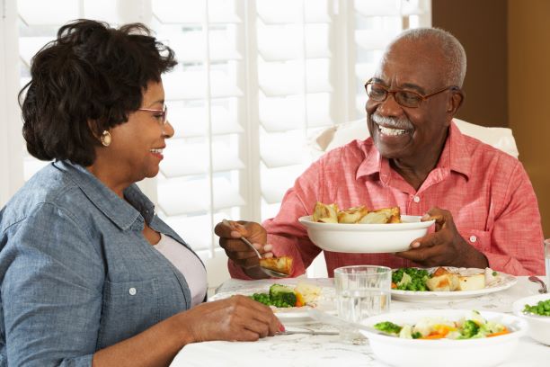 Un hombre mayor le sirve comida de un recipiente a una mujer sentada a su lado en una mesa de comedor.