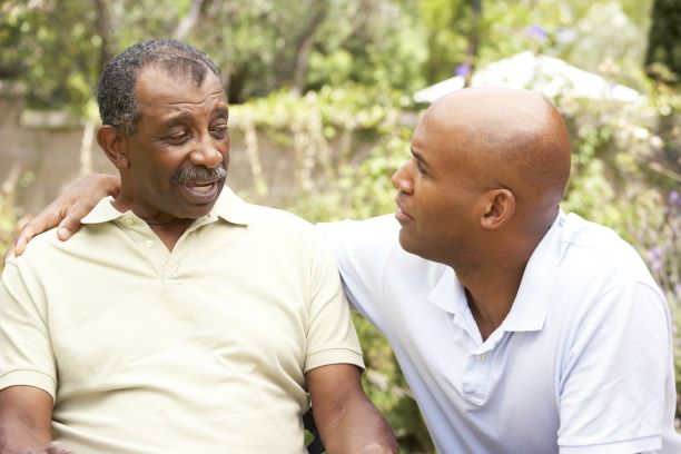 Un hombre mayor está sentado mientras habla con un hombre más joven que descansa su brazo sobre el hombro del hombre mayor.
