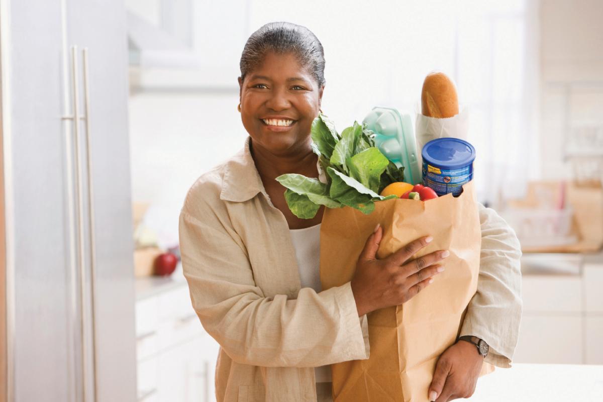 A smiling woman holding a grocery bag of food in her kitchen.