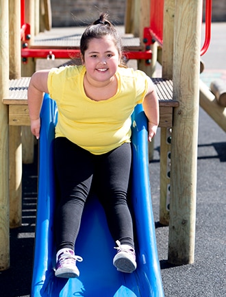 A young girl with overweight or obesity who is sliding down a slide.