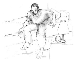 Male getting out of his bed, next to him a bed-side table with lamp.