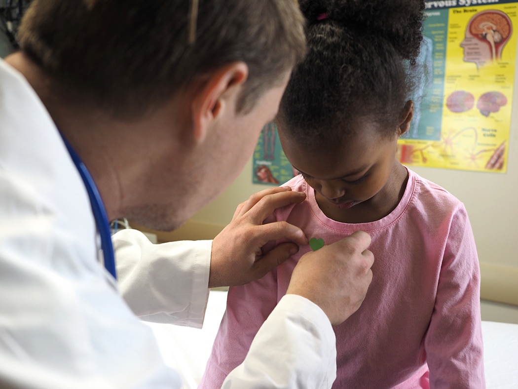 Health care professional places a sticker on the shirt of a little girl who is sitting on an examining table.