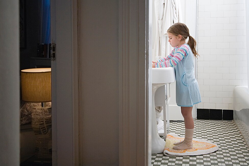 View from a hallway shows a girl washing her hands in a bathroom next to her bedroom.
