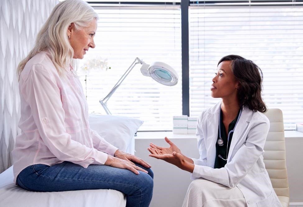 Una paciente femenina en el consultorio hablando con una profesional de atención médica en una camilla de examinación.