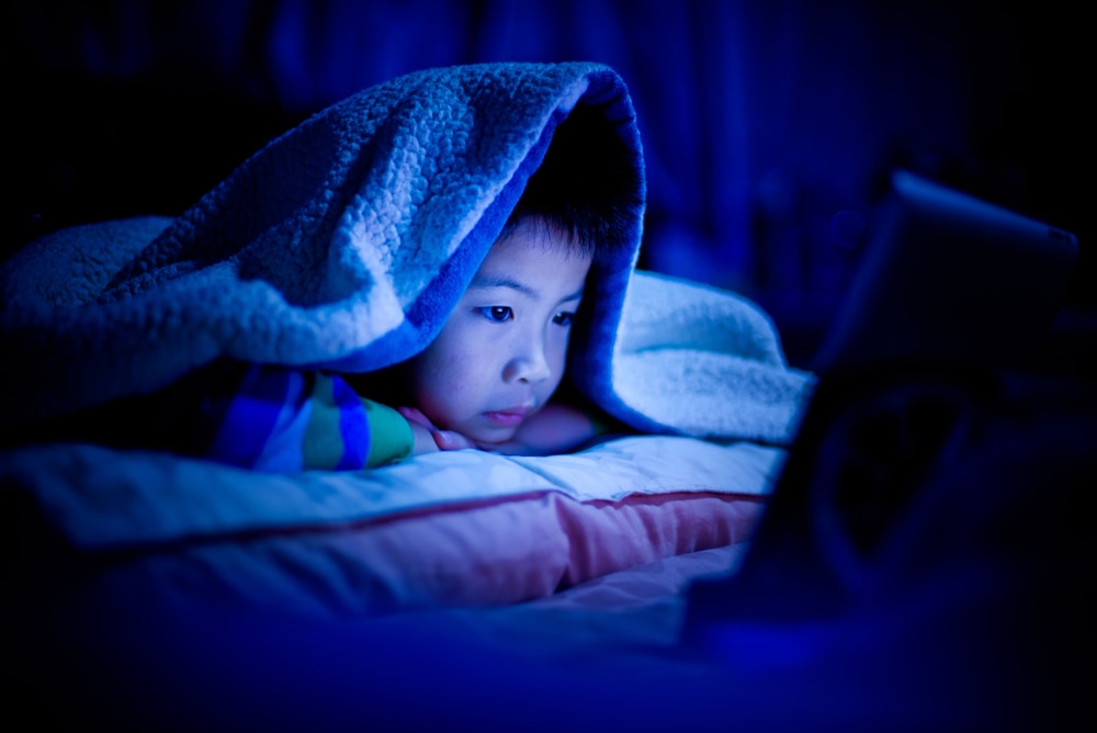 Boy lying on his stomach in bed, under the covers, with the lights turned off, looks at a screen that emits blue light.