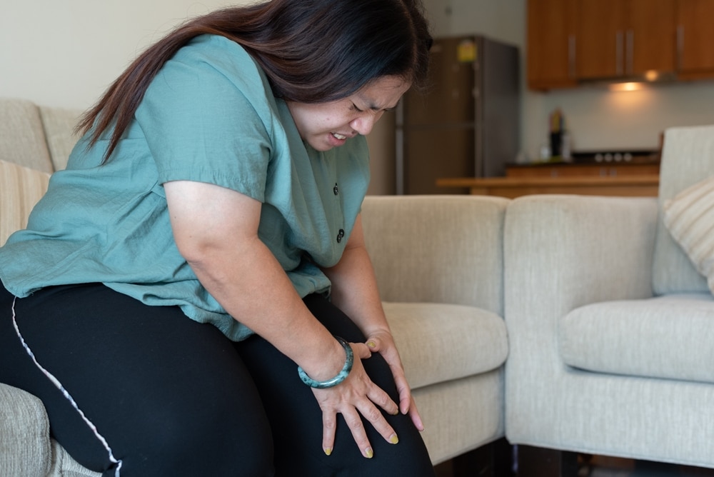 A woman with overweight sitting on a sofa holding her knee in pain.