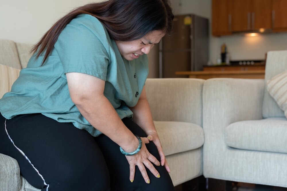 Una mujer con sobrepeso está sentada en un sofá, se toca la rodilla y tiene cara de dolor.