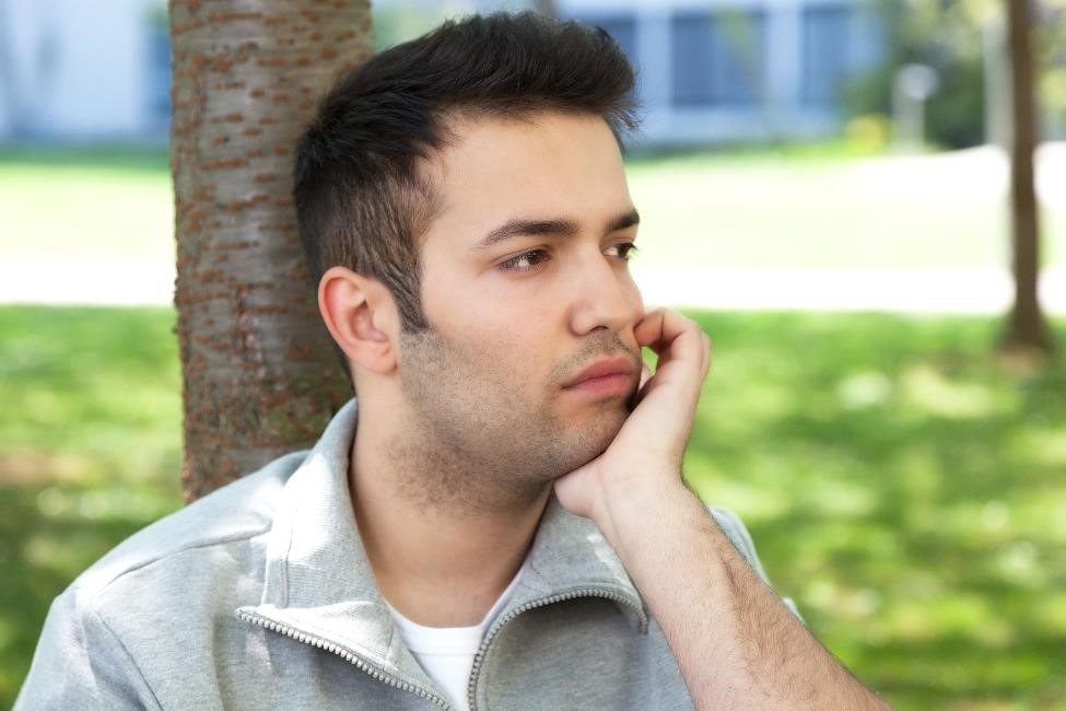A young man sits in a park with his hand on his chin.