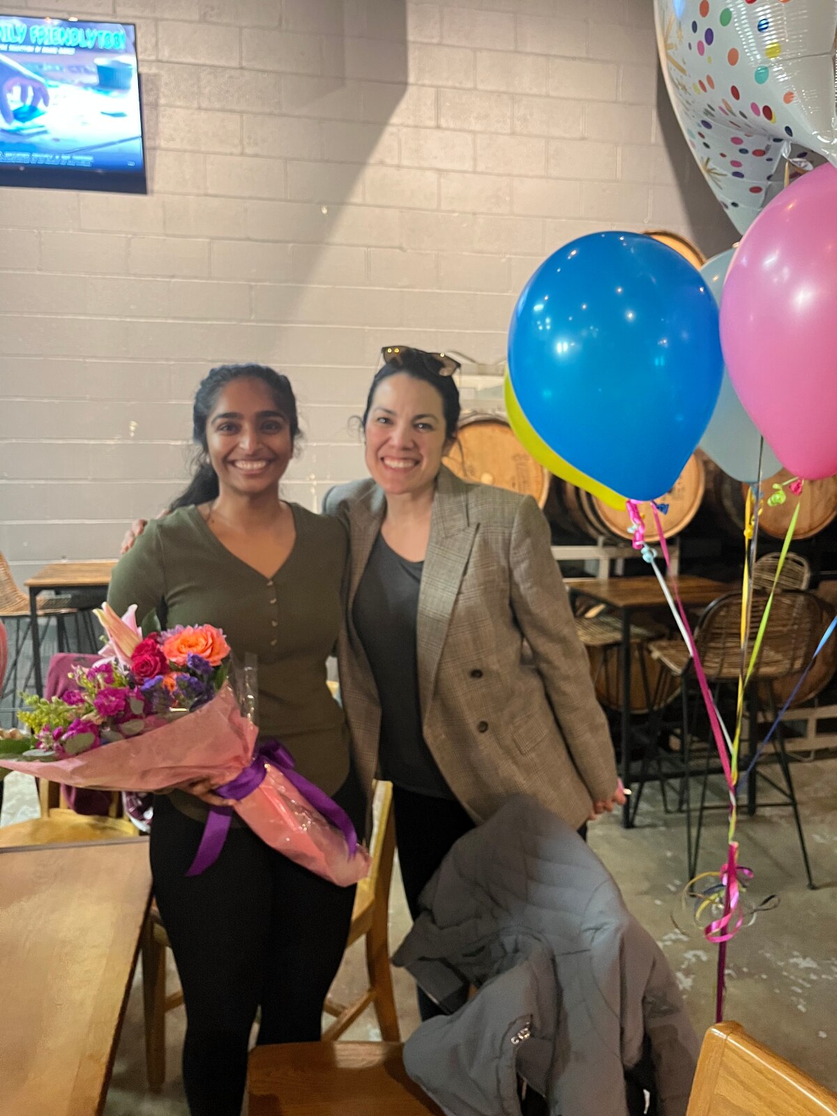 Two women smiling next to party balloons. One is holding a bouquet of flowers.