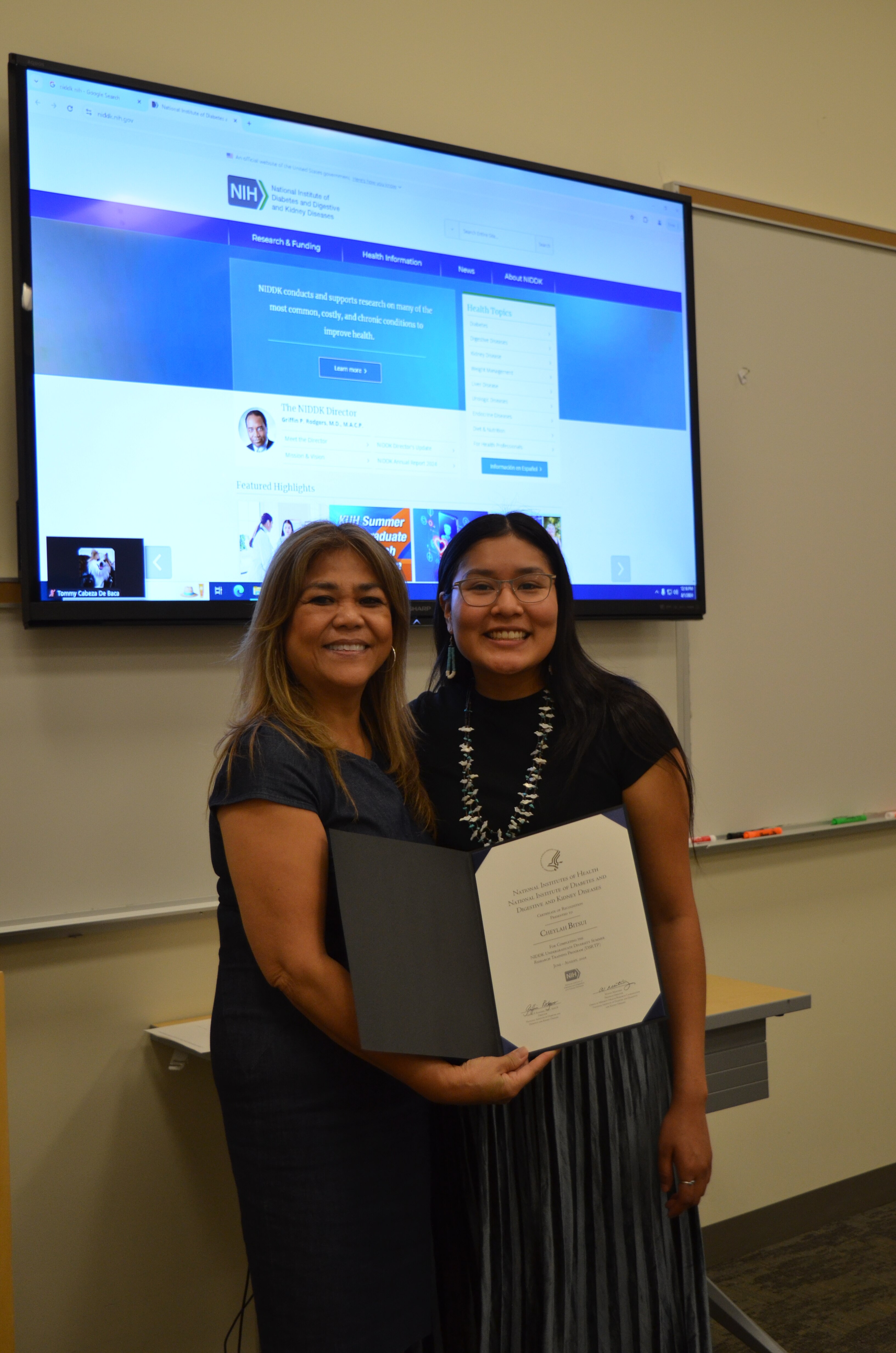 Two women holding a certificate and smiling in front of a screen showing an online meeting.